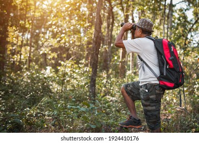 Old Man With Backpack Looking At Binoculars In The Forest. Elderly Man Hiking On Vacation