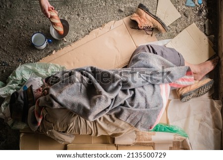 Old man beggar homeless wears sweater with blanket sleeping on cardboard at shelter with bowl for asks donate food and money. Hand of people kind give bread for poor poverty hungry and sad on street.