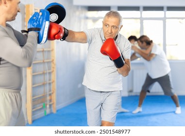 Old man in boxing gloves practices punches with man in mitts in gym, man is practicing sparring with gyu in background - Powered by Shutterstock