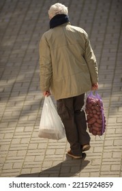 An Old Man Carries A Heavy Bag And A Sack Of Potatoes In His Hands