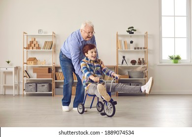 Old Man Is Child Again. Healthy Senior Man Enjoying Free Time And Playing Childish Games Together With Teen Grandson. Happy Excited Little Boy Riding Tricycle And Having Fun With Grandfather At Home