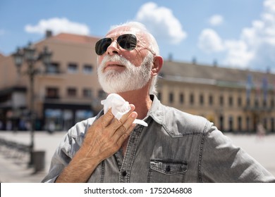 Old Man Cleaning Neck And Sweat With Wet Wipes On Street On Hot Summer Day, Blurred Urban City Background