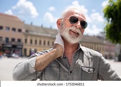 Old Man Cleaning Neck And Sweat With Wet Wipes On Street On Hot Summer Day, Blurred Urban City Background