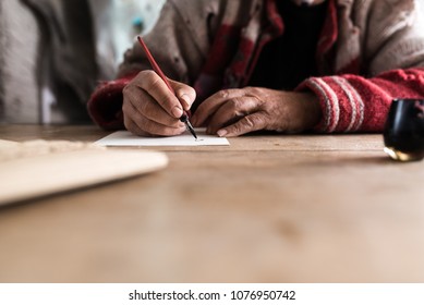 Old Man With Dirty Hands Writing A Letter Using A Nib Pen And Ink Or Pigment From A Pot In A Low Angle View Across A Wooden Table.