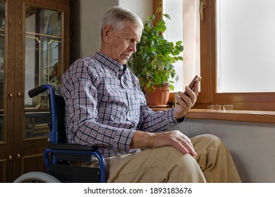 An Old Man With A Disability In A Wheelchair Sitting At Home, Looking At Mobile Phone, Use The Internet.