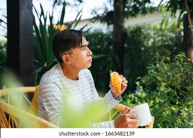 Old Man Eating Sausage Croissant And Holding The Mug In The Backyard Of His House.