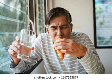 Old Man Eating Sausage Croissant While Hold A Glass Of Drinking Water In Another Hand.