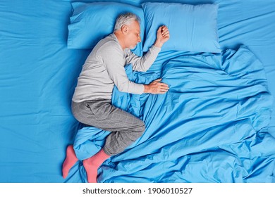 Old man enjoys healthy sleeping feels comfortable on bed lying at soft fresh linen wears pajama and socks fell asleep immediately after hard working day. Grey haired pensioner napping in morning