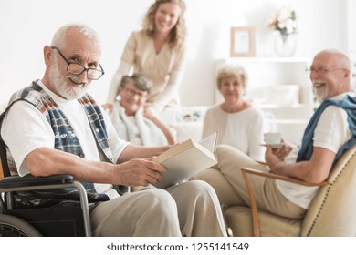 Old Man With Glasses Sitting On Wheelchair Reading A Book