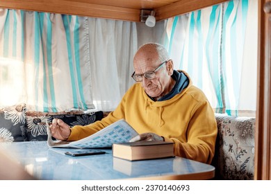 Old man in glasses sitting at table inside of recreational vehicle and solving crossword in newspaper - Powered by Shutterstock