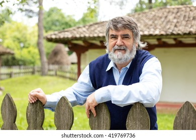 Old Man With Gray Hair And Beard Standing In Front Of His Old House Behind An Wooden Fence