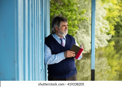 Old Man With Gray Hair Standing On A Porch Next To A Lake With A Book