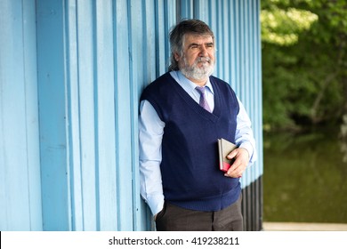 Old Man With Gray Hair Standing On A Porch Next To A Lake With A Book