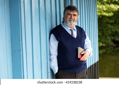 Old Man With Gray Hair Standing On A Porch Next To A Lake With A Book