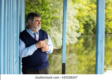 Old Man With Gray Hair Standing On A Porch Next To A Lake With A Book