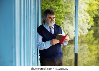 Old Man With Gray Hair Standing On A Porch Next To A Lake With A Book