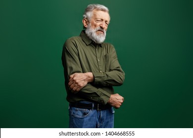 Old Man In Green T-shirt And Jeans With Closed Eyes Holding His Elbow In Pain In The Studio With Green Wall, Help Me, I've Broken My Arm