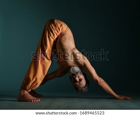 Old man with grey beard doing yoga, pilates, fitness training, stretching exercise, asana or balance workout on floor over dark blue background. Healthy lifestyle concept