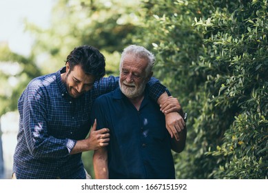 The Old Man And His Son Are Walking In The Park. A Man Hugs His Elderly Father. They Are Happy And Smiling