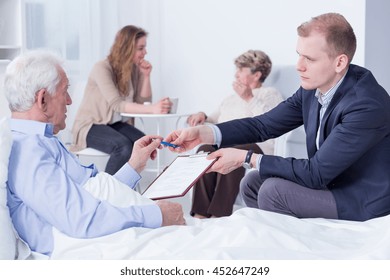 Old Man In A Hospital Bed Being Handed Documents To Sign By A Lawyer Sitting Next To Him