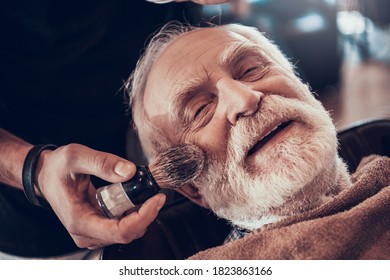 Old Man Looking At Camera While Applying Shaving Gel In Barbershop. The Old Grandfather Sits In A Barber Chair While Shaving And Smiles.