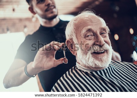 An old man looks at the camera while the master cuts his hair in a barbershop. An adult sidoy dede looks at the camera while he gets a stylish haircut. 