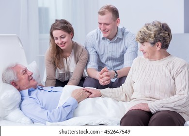 Old man lying in a white hospital bed, surrounded by his smiling relatives