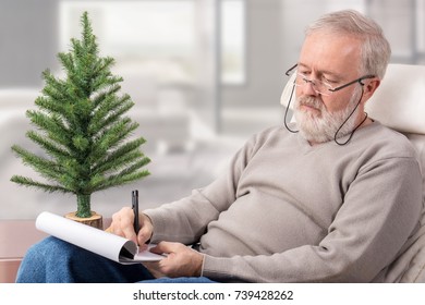 Old Man Making A Shopping List With A Pen On Paper Next To An Undecorated Fir Tree For Christmas Holidays