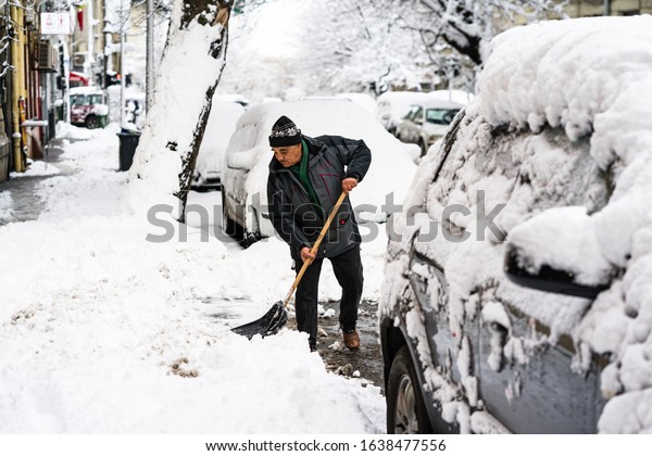 Old man, male in\
winter coat cleaning, shoveling driveway, street from covered\
falling snow with shovel, residential houses, cars parked on road\
in Bucharest, Romania 2020