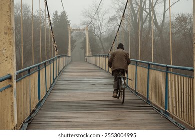 An old man on an old bicycle rides over a boardwalk, suspension bridge. Autumn foggy weather, the dreary atmosphere of late fall - Powered by Shutterstock