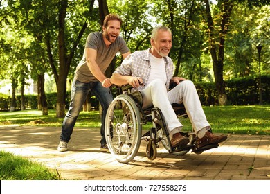 The old man on a wheelchair and his adult son are walking in the park. A man is carrying his father, they are having fun and they are laughing
