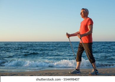 Old man in orange shirt black shorts making nordic walking on beach. Senior man doing sports on vacation on sea coast