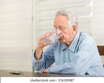 Old Man In Pajamas Drinking Glass Of Water And Have Pills Blister On Table