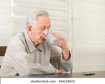 Old Man In Pajamas Drinking Glass Of Water And Have Pills Blister On Table