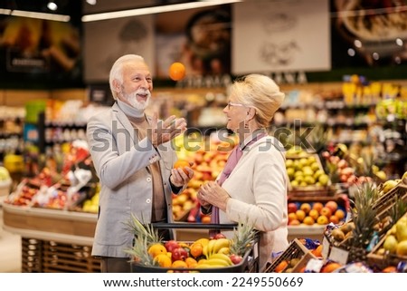 An old man is playing with fruits while his wife watching and smiling at him. A senior couple at the supermarket.