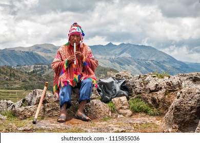 The old man Quechua dressed in a colored poncho and a cap of Chullo, sits on the rocks with a view of the mountains in the background and plays on the musical instrument of Quena. Cusco, Peru.