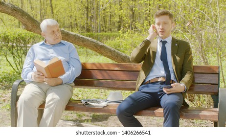 Old Man Is Reading Book In Th Park And Businessman Is Disturbing It With Loud Conversation On Phone With Audio Headset