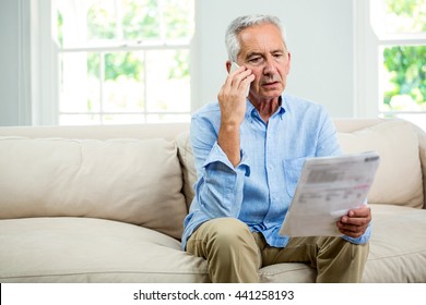 Old man reading document while talking on phone in living room at home
