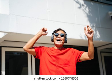 Old Man In Red T-shirt Enjoy The Music From Headphone And Dance In Front Of His House In Sunny Day.