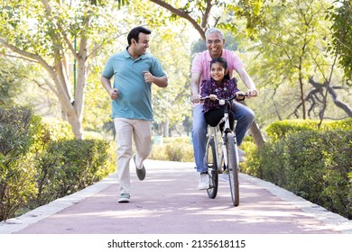 Old man riding bicycle with granddaughter while son running at park
