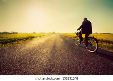 Old man riding a bike on asphalt road towards the sunny sunset sky