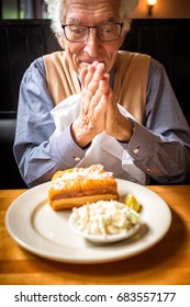 Old Man Rubs His Hands In Anticipation Of Eating The Food In Front Of Him. Restaurant Setting. Happy Man Wearing A Nice Shirt And Sweater. He's Hungry And Excited About The Lobster Roll On His Plate.