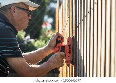 Old Man Sanding Wood Fence By Sander. Senior Man Using Work Tool. Renovation Of Wooden Picket Fence At Backyard