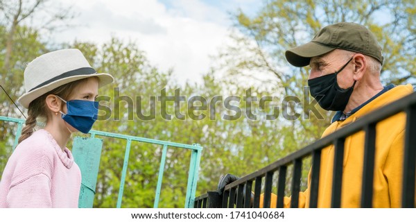 old man and\
schoolgirl in disposable masks stand at distance divided by village\
metal fence against garden\
closeup
