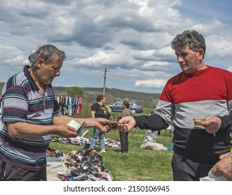 An Old Man Selling Shoes At The Fair Reaches Out With The Rest Of The Money To The Customer. Poor Fair With Worn Clothes And Shoes. Romania, Mehedinti. April, 26, 2022