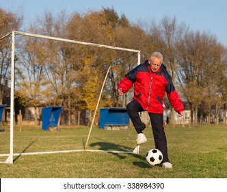 Old Man In Seventies Kicking A Soccer Ball On Playground With Goal Behind Him