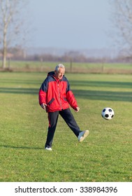 Old Man In Seventies Kicking A Soccer Ball On The Grass Field