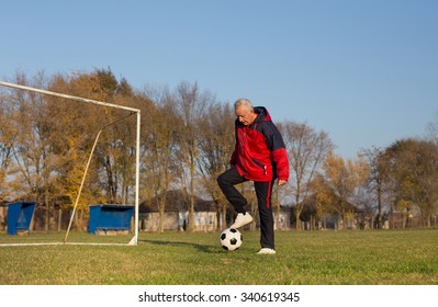 Old Man In Seventies Kicking A Soccer Ball On Playground With Goal Behind Him