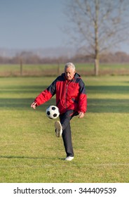 Old Man In Seventies Kicking A Soccer Ball On The Grass Field