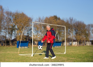 Old Man In Seventies Kicking A Soccer Ball On Playground With Goal Behind Him